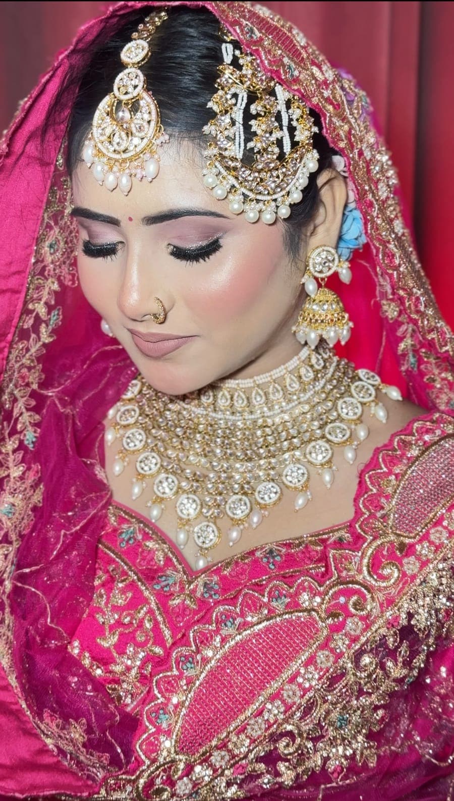 Bride in a pink lehenga and veil with layered gold jewellery, looking down