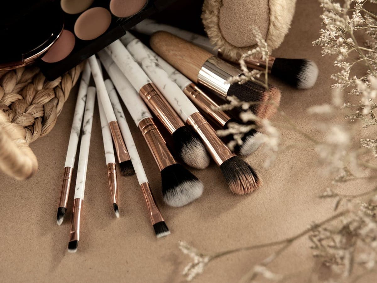 Marble-handled makeup brushes with rose-gold detail on a champagne surface beside dried flowers